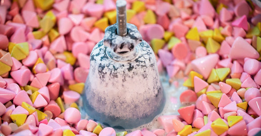 Industrial tumbling machine full of pink and yellow plastic polishing media. A white cleaning solution is visible underneath the plastic pieces.