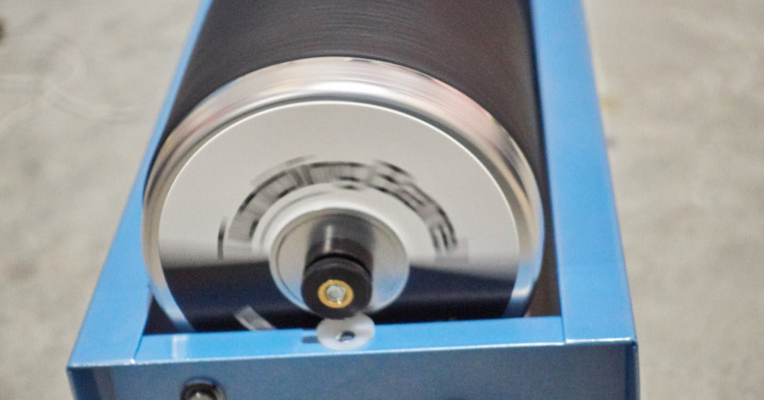 A black rotary tumbler barrel spinning in a brightly lit industrial facility. The barrel is attached to a blue machine.