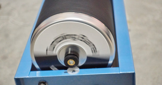 A black rotary tumbler barrel spinning in a brightly lit industrial facility. The barrel is attached to a blue machine.