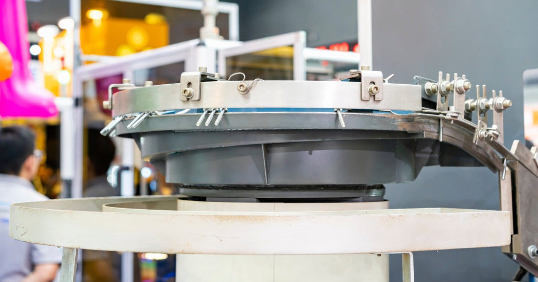 A small gray vibratory finishing bowl installed in an industrial facility. An employee nearby looks at another machine.
