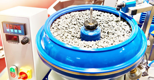 A blue vibratory finishing machine full of gray media. A nearby faucet dumps water into the vibratory bowl.